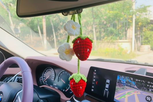 a crocheted strawberry and flower keychain hanging from rear view mirror in a car car has light pink front interior