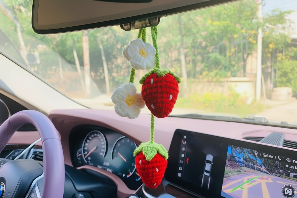 a crocheted strawberry and flower keychain hanging from rear view mirror in a car car has light pink front interior

